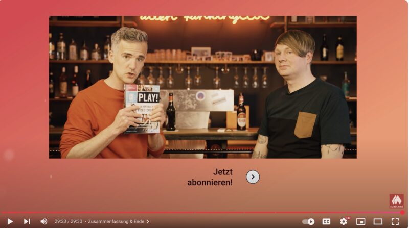 Zwei Männer stehen in einer Bar-Szenerie mit beleuchtetem Regal im Hintergrund. Der Mann links hält die Zeitschrift „PLAY! Das Handbuch für Video-Creator“ in die Kamera und spricht, während der Mann rechts ruhig in die Kamera schaut. Im unteren Bereich erscheint der Hinweis „Jetzt abonnieren!“ mit einem Pfeilsymbol.