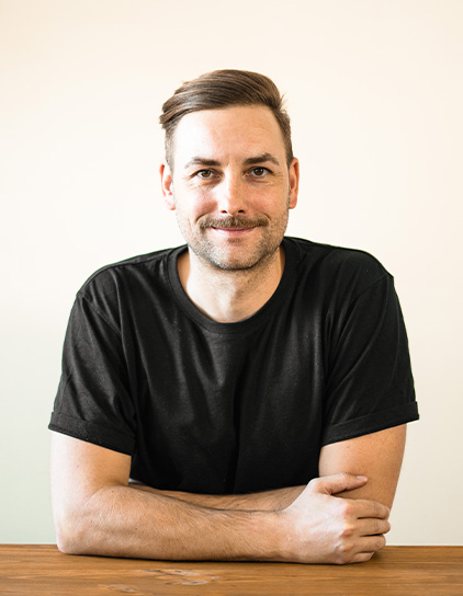 Bild von Alex Welk, Mitarbeiter bei Morefire, in einem schwarzen T-Shirt sitzend