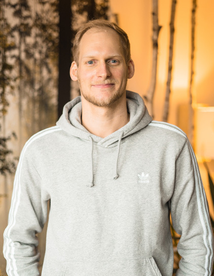 Lars Woestmann, Mitarbeiter im grauen Adidas-Hoodie, stehend im Büro vor einer Wand mit Baumdekor