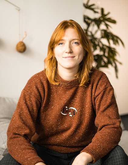 Leah Gries, Mitarbeiterin mit rotem Pullover, sitzt im Büro vor einer grünen Pflanze