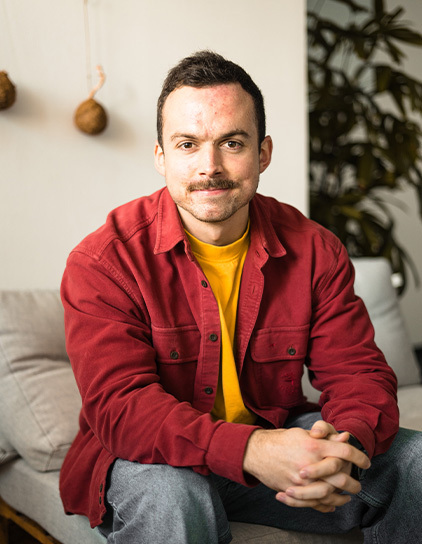 Leon Krieger trägt ein rotes Hemd und ein gelbes T-Shirt, sitzt auf einem Sofa