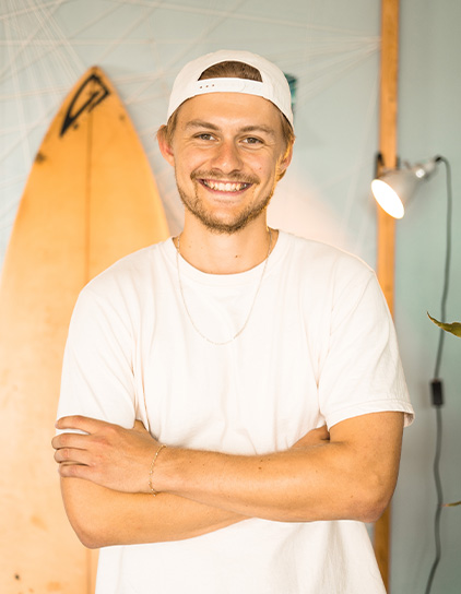Marvin Foppe lächelt in einem weißen T-Shirt und einer Kappe vor einem Surfbrett bei MoreFire
