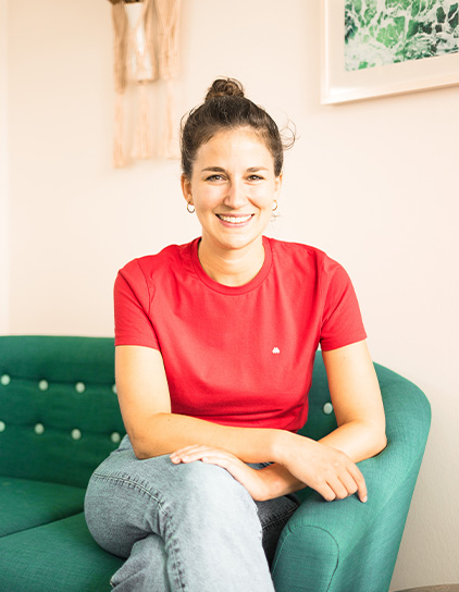 Pia Dederichs in einem roten T-Shirt, sitzt auf einem grünen Sofa und lächelt in die Kamera