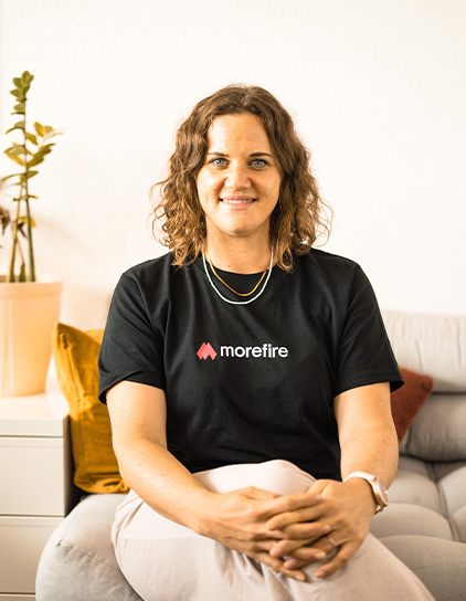 Ramona Schäfer in einem schwarzen Morefire-T-Shirt, sitzt auf einem Sofa und lächelt in die Kamera
