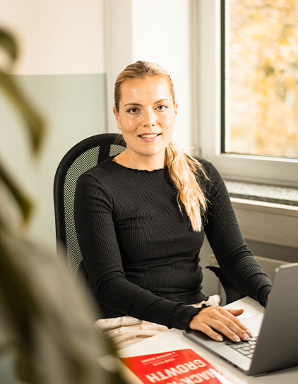 Rebecca Barth in einem schwarzen Pullover, sitzt am Schreibtisch und lächelt in die Kamera