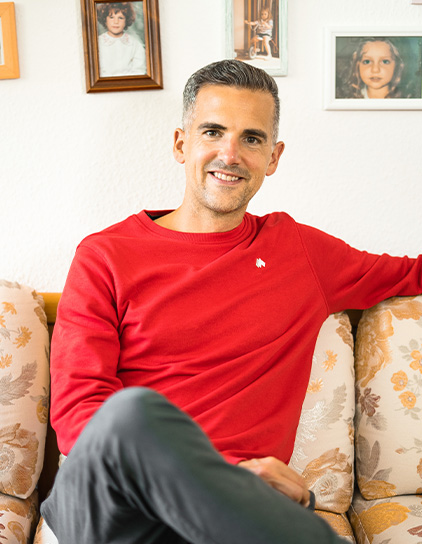 Robin Heintze in einem roten T-Shirt, sitzt auf einem Sofa und lächelt in die Kamera"