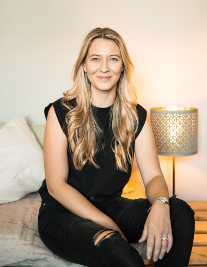 Sarah Glittenberg in einem schwarzen T-Shirt, sitzt auf einem Bett und lächelt in die Kamera