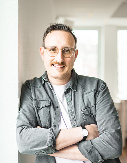 Sebastian Carlsson in einem grauen Hemd, lehnt an einer Wand und lächelt in die Kamera