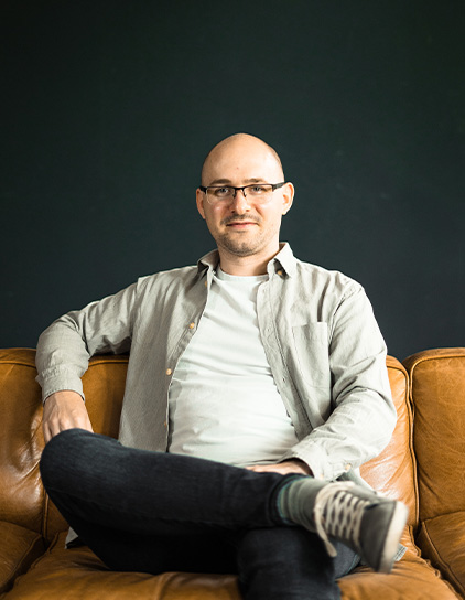 Thomas Kaiser in einem grauen Hemd, sitzt auf einem Ledersofa und lächelt in die Kamera