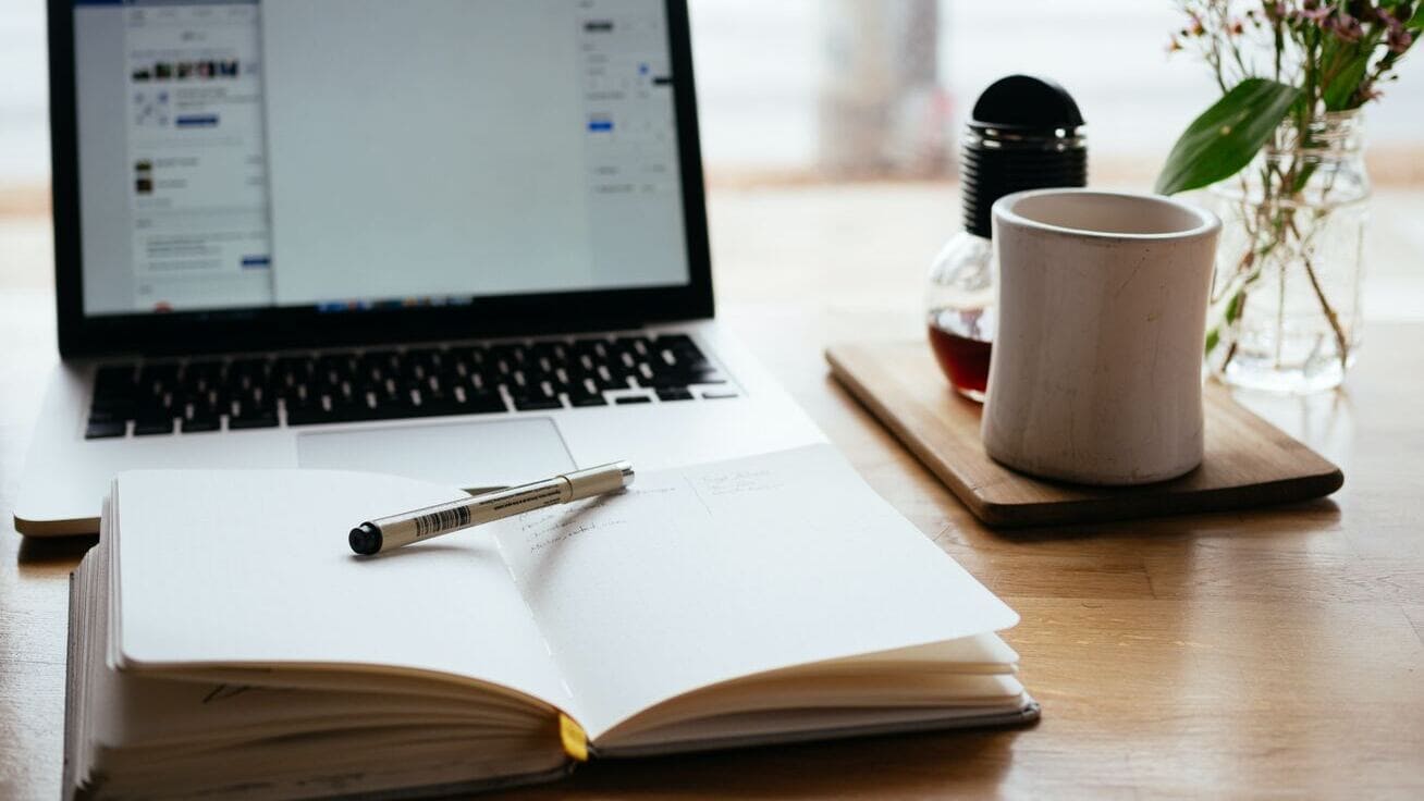 Laptop mit offenem Notizbuch und Kaffeetasse auf einem Schreibtisch